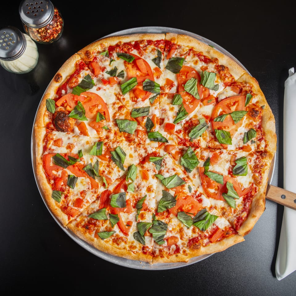Margherita pizza with melted mozzarella, tomato slices, and fresh basil on a metal tray beside seasonings and a napkin.