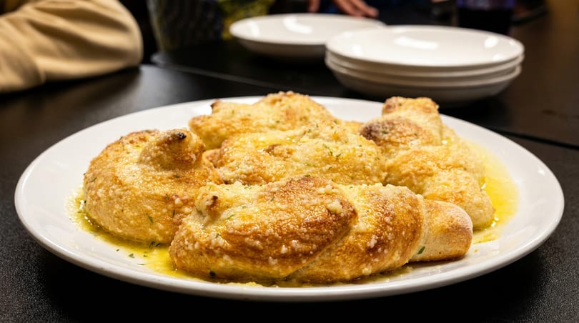 Golden-brown garlic knots drenched in butter, cheese, and herbs on a white plate.
