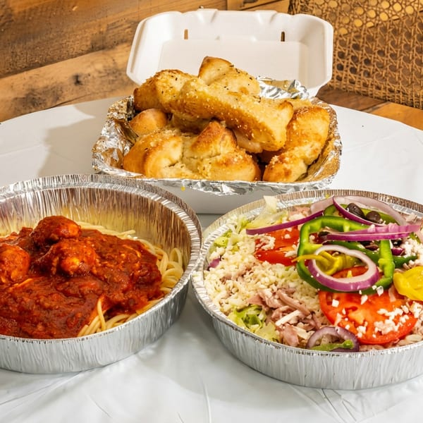 Spaghetti with meatballs, a large salad, and garlic knots in takeout containers on a white table.