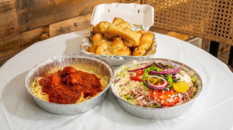 Spaghetti and meatballs, a large salad, and garlic knots in takeout containers on a white table.