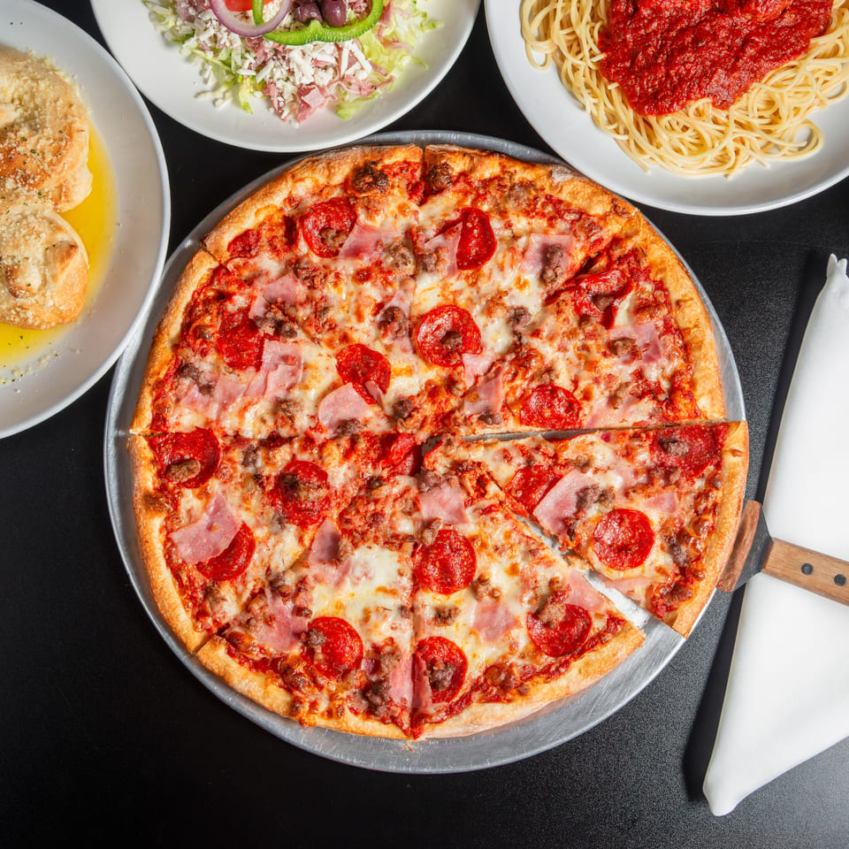 Overhead view of a pepperoni, ham, and sausage pizza surrounded by garlic bread, salad, and spaghetti with marinara sauce.