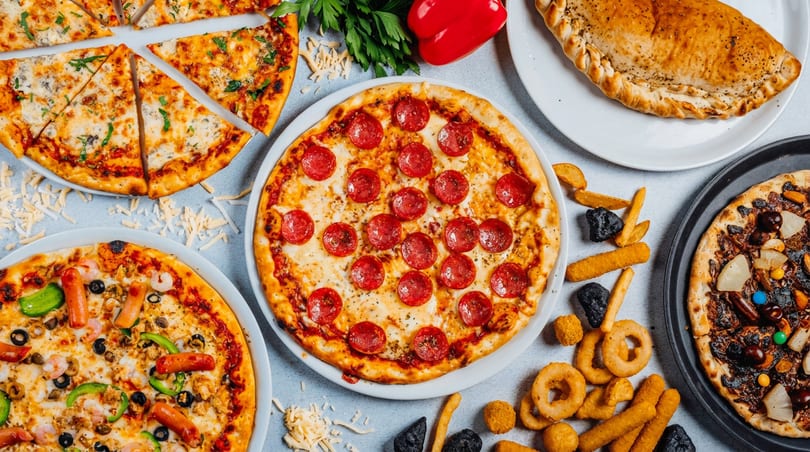 A table filled with diverse pizzas, a calzone, and fried appetizers like onion rings and fries.