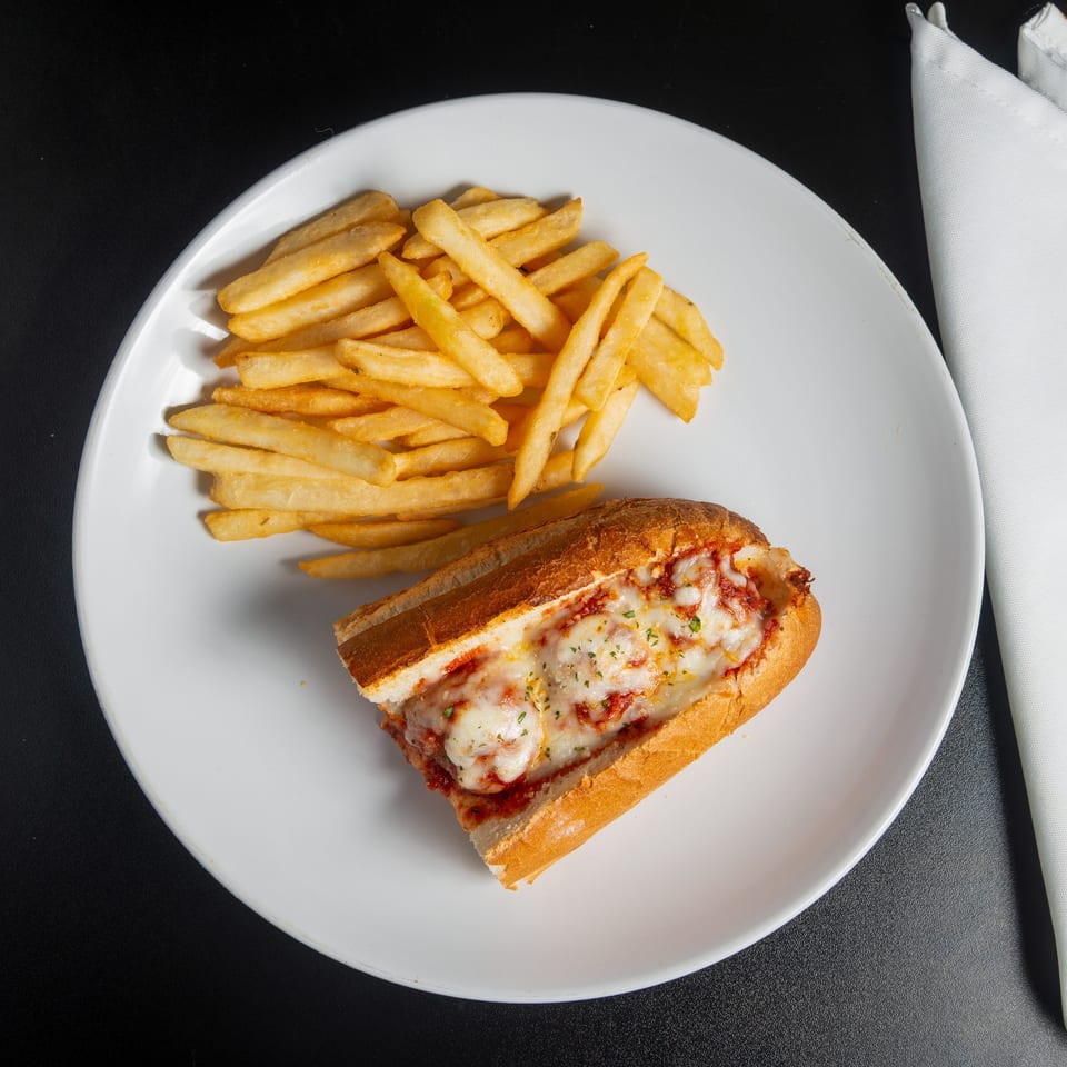 A white plate holds a cheesy meatball sub with marinara and parsley beside golden french fries on a black table.