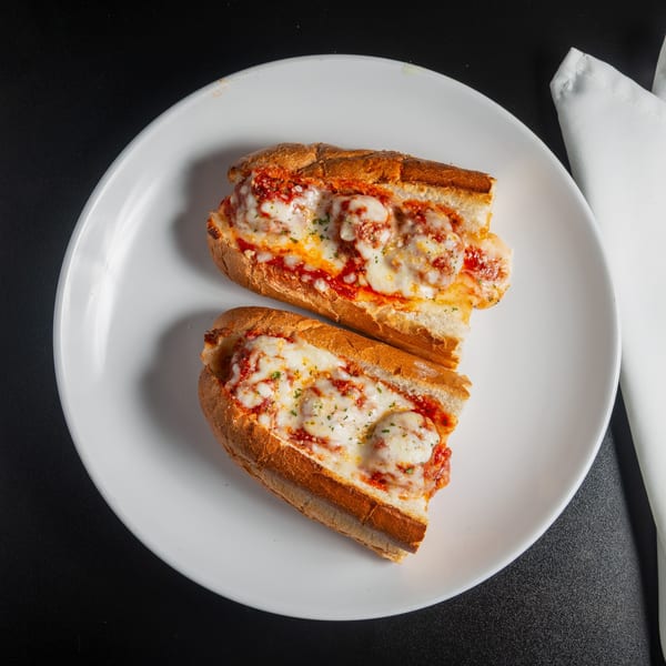 Two meatball subs topped with marinara, melted cheese, and herbs on a white plate beside folded napkins.