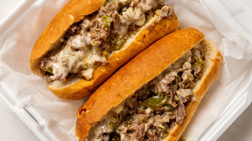 Two cheesesteak sandwiches with green peppers and melted cheese in a white takeout container.