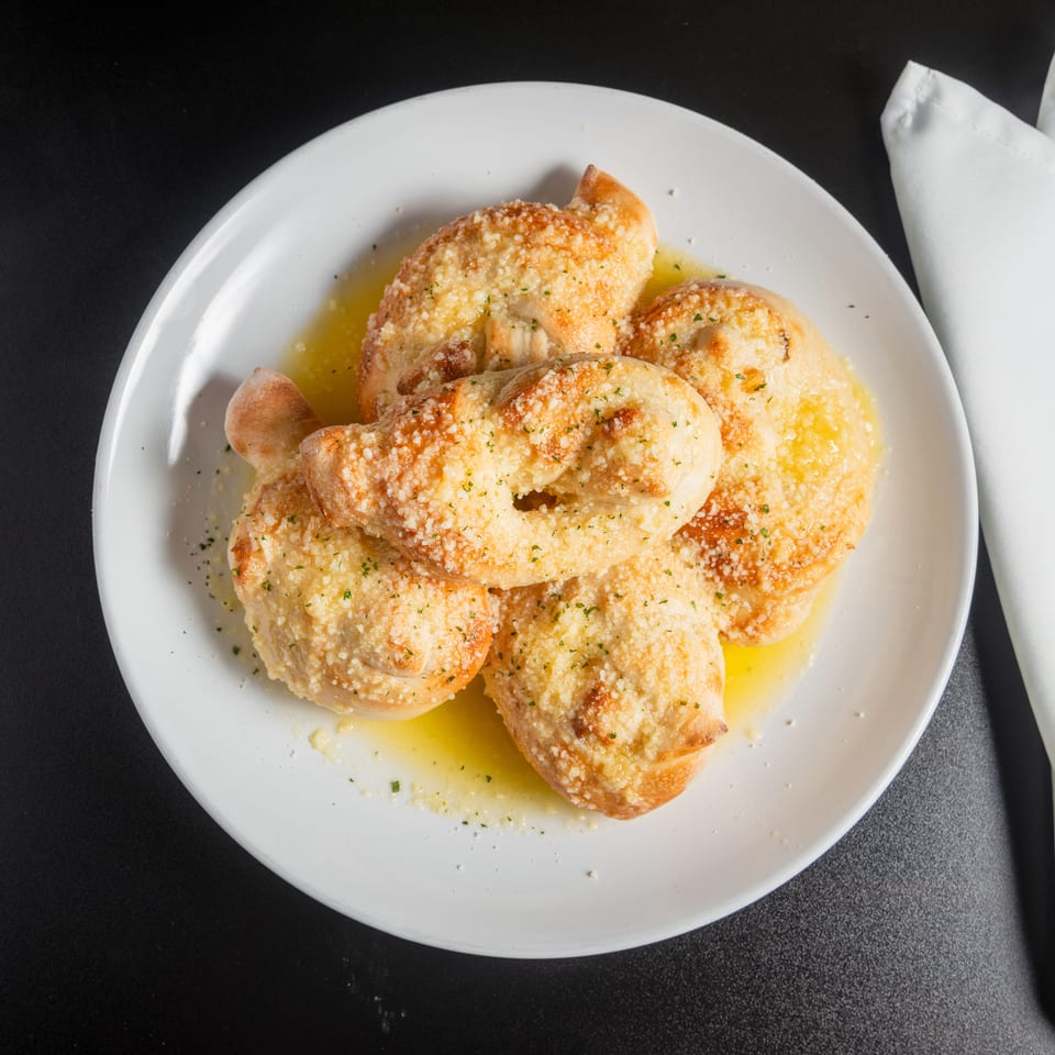 Golden garlic knots topped with Parmesan and herbs, served in melted butter on a white plate beside a napkin.