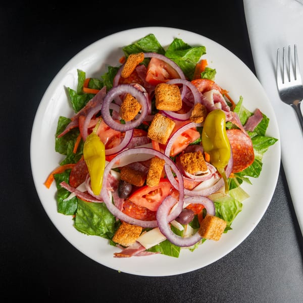Antipasto salad with romaine lettuce, tomatoes, onions, cured meats, pepperoncini, olives, cheese, and croutons on a white plate.