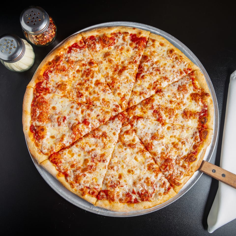 Overhead view of a large sliced cheese pizza on a metal tray with red pepper flakes, seasonings, and a napkin.
